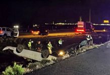 Nighttime highway crash scene with an overturned car on a median barrier and emergency responders nearby, lights flashing in the distance.