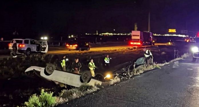 80 double fatal Nighttime highway crash scene with an overturned car on a median barrier and emergency responders nearby, lights flashing in the distance.