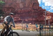 Adult cyclist in the foreground rides a mountain bike on a sunny road, with two children biking behind near red rock cliffs.