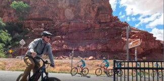 Adult cyclist in the foreground rides a mountain bike on a sunny road, with two children biking behind near red rock cliffs.