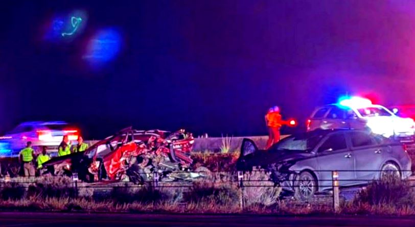 Nighttime highway crash scene with several wrecked cars, emergency responders in reflective vests, and flashing blue and red police lights.