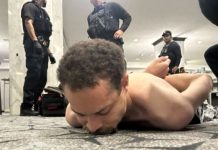 Police officers in tactical gear stand around a shirtless man lying face down on a patterned carpet in a bright indoor space, as if restraining him.