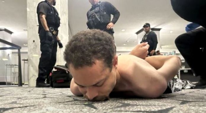 Police officers in tactical gear stand around a shirtless man lying face down on a patterned carpet in a bright indoor space, as if restraining him.