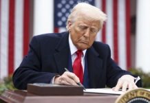 A man in a dark suit and red tie signs a document at a desk outdoors, with American flags in the background.