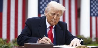 A man in a dark suit and red tie signs a document at a desk outdoors, with American flags in the background.