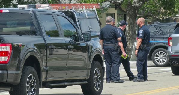 Three police officers stand in a parking lot beside dark SUVs, talking to each other, with one holding a yellow object.