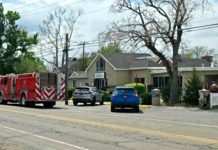 Red fire trucks parked on a suburban street in front of a beige, single-story building; a blue car and a gray SUV in front, with leafless trees and power lines overhead.