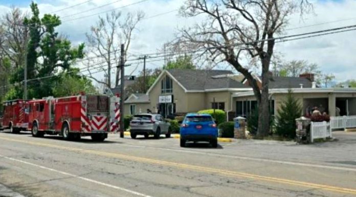 Red fire trucks parked on a suburban street in front of a beige, single-story building; a blue car and a gray SUV in front, with leafless trees and power lines overhead.