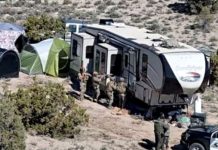Military personnel stand near a large RV and several tents set up on a desert hillside, with a sheriff's vehicle nearby.