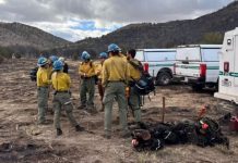 Firefighters in yellow shirts and blue helmets stand in a circle on a dry field, with gear and emergency vehicles nearby.
