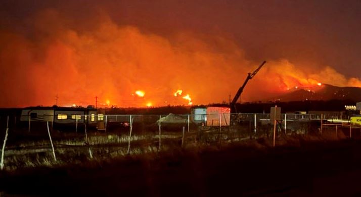 Nighttime wildfire burning across a field with bright orange flames and thick smoke, a fence and parked vehicles visible in the foreground.