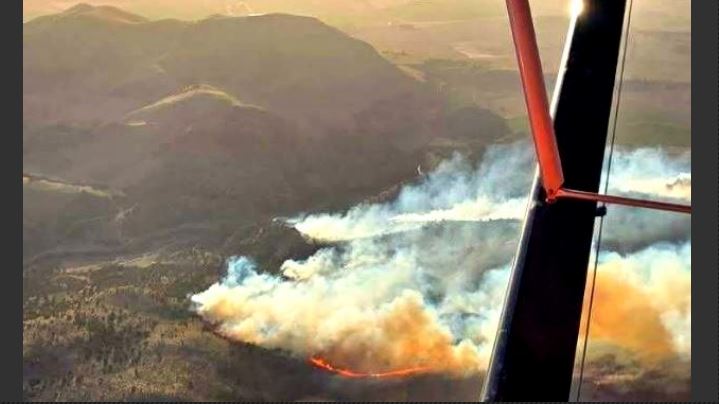Aerial view of a forest fire with flames and smoke below, seen from a aircraft wing over hilly terrain.