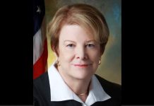 Formal portrait of a middle-aged woman with short blonde hair, wearing a black blazer and white blouse, with an American flag in the background.