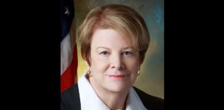 Formal portrait of a middle-aged woman with short blonde hair, wearing a black blazer and white blouse, with an American flag in the background.