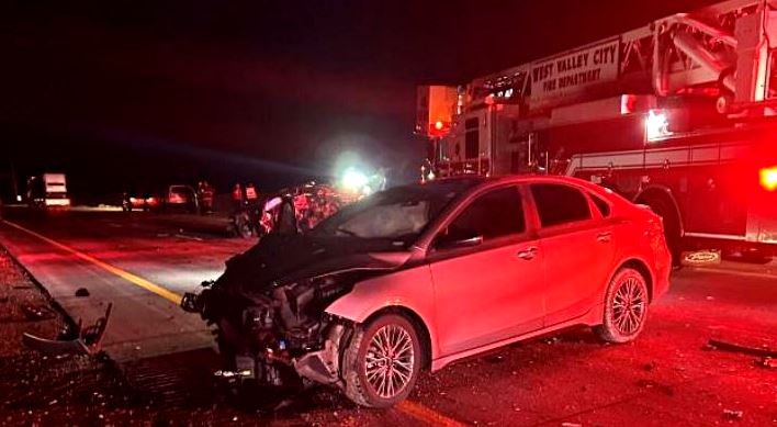 Red sedan with severe front-end damage on a highway at night, emergency lights and a fire truck in the background.