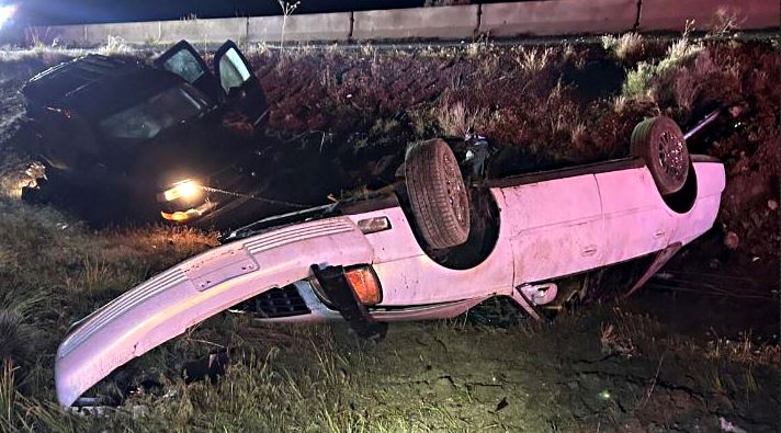White sedan overturned on its roof in a ditch beside a grassy embankment, headlights on, nighttime scene