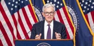 Older man in a dark suit and purple tie speaks at a podium, with US flags and a Board of Governors seal behind him, microphones visible