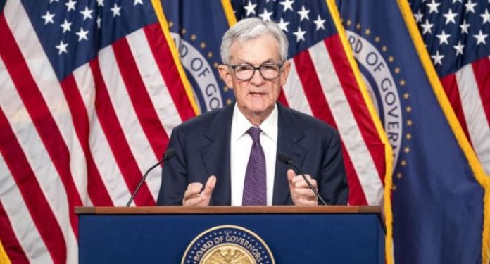 Older man in a dark suit and purple tie speaks at a podium, with US flags and a Board of Governors seal behind him, microphones visible