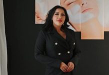 Woman in a black double-breasted blazer stands against a dark wall with peach-colored portrait panels in the background.