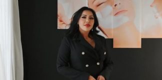 Woman in a black double-breasted blazer stands against a dark wall with peach-colored portrait panels in the background.