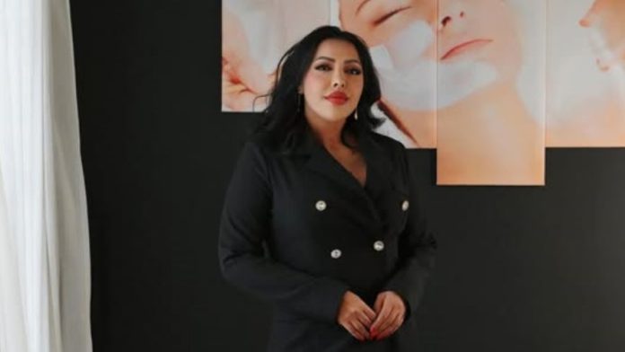 Woman in a black double-breasted blazer stands against a dark wall with peach-colored portrait panels in the background.
