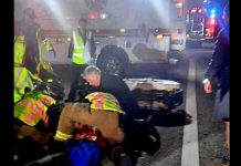 Emergency responders in reflective vests aid a person on the ground at night on a street, with ambulance lights in the background.