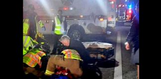 Emergency responders in reflective vests aid a person on the ground at night on a street, with ambulance lights in the background.