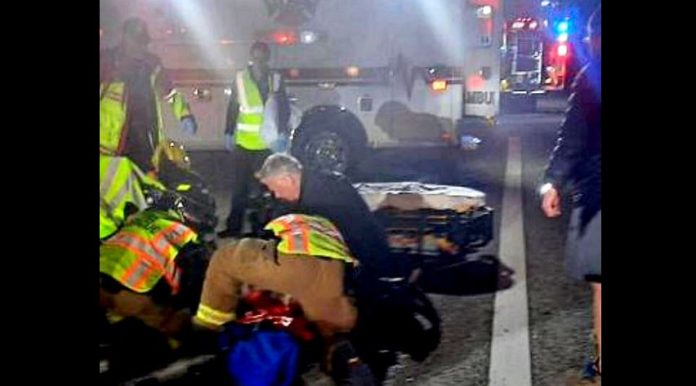 Emergency responders in reflective vests aid a person on the ground at night on a street, with ambulance lights in the background.