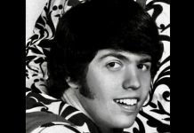 Black-and-white portrait of a young man with dark hair, smiling, against a bold floral-patterned background.
