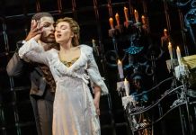 Masked man in a dark suit holds a woman in a white lace gown on a candlelit stage with ornate ironwork behind them.
