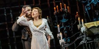 Masked man in a dark suit holds a woman in a white lace gown on a candlelit stage with ornate ironwork behind them.