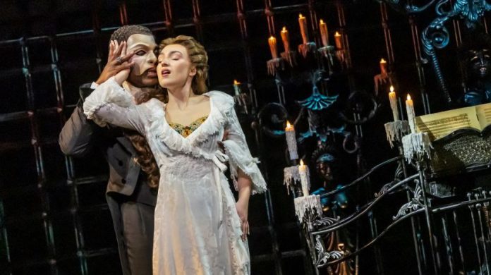 Masked man in a dark suit holds a woman in a white lace gown on a candlelit stage with ornate ironwork behind them.