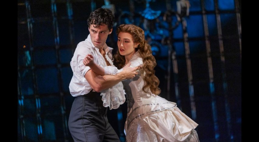 Two dancers in period costumes perform a ballroom dance on a dark stage, the man guiding the woman with a tense, protective hold.