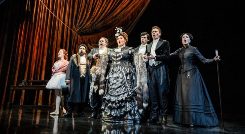 Group of opera singers in Victorian-era costumes standing on stage for a curtain call, with a gold drapery backdrop behind them.