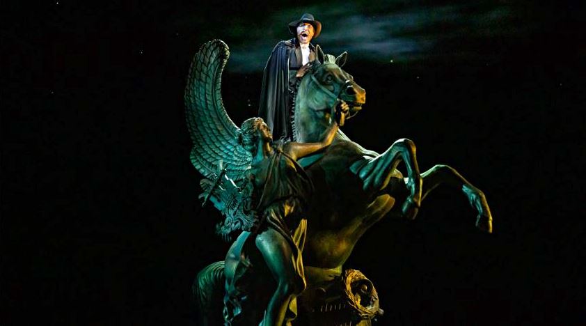 Statue of a rider in a hat and cloak on a winged horse, illuminated against a dark night background.