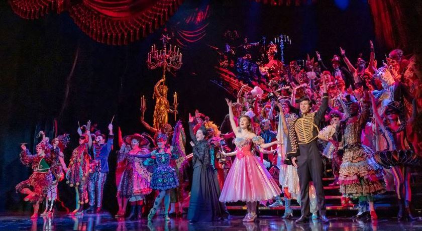 Large cast in colorful, period-style costumes performs on a lit theater stage, arms raised in a finale pose.