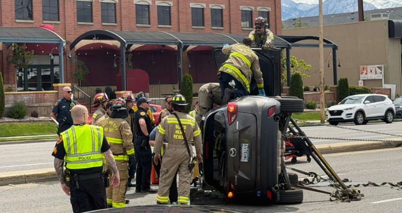 Firefighters and police surrounding a gray SUV flipped on its side during a rescue on a city street
