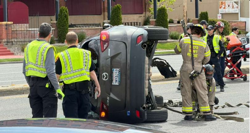 Gray SUV overturned on its side in the middle of a city street, surrounded by responders in bright yellow vests and firefighters in tan turnout gear, with a tow strap on the road.