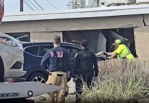 Emergency responders and police stand near a dark blue car wedged into a damaged storefront after a crash at a street corner.