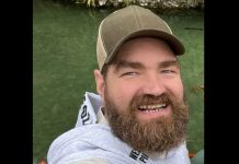 Close-up of a man with a full beard wearing a brown cap, smiling at the camera with a river in the background.