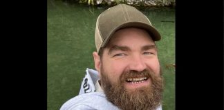 Close-up of a man with a full beard wearing a brown cap, smiling at the camera with a river in the background.