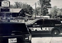 Black-and-white photo of several police SUVs parked in a lot outside a building with a sign for Fresh Start Community Health Center and Optum nearby.