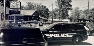 Black-and-white photo of several police SUVs parked in a lot outside a building with a sign for Fresh Start Community Health Center and Optum nearby.
