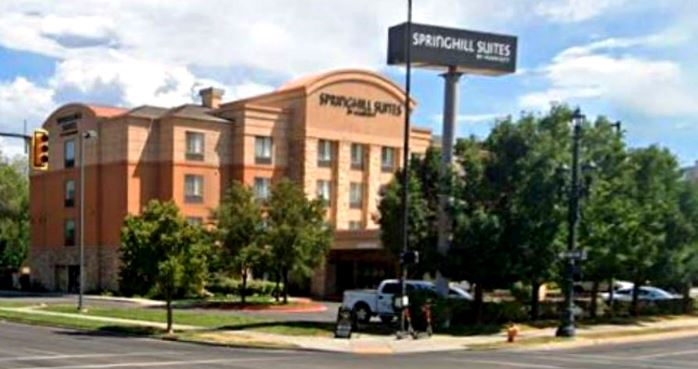Exterior of a SpringHill Suites hotel at a tree lined street corner with a tall sign and parked cars nearby