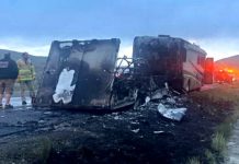Burned-out large truck on the highway shoulder, charred interior exposed, with firefighters and an emergency vehicle with flashing lights in the background.