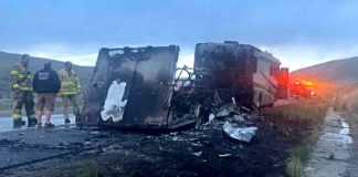 Burned-out large truck on the highway shoulder, charred interior exposed, with firefighters and an emergency vehicle with flashing lights in the background.