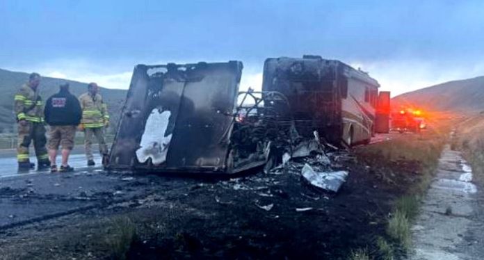 Burned-out large truck on the highway shoulder, charred interior exposed, with firefighters and an emergency vehicle with flashing lights in the background.