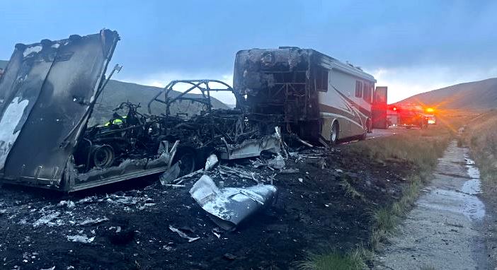 Wrecked bus trailer and burned-out car debris along a rural roadside, emergency vehicles with flashing lights in the distance.