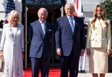 Two well-dressed couples stand on a red carpet outside a white building with flags, smiling for the camera.