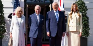 Two well-dressed couples stand on a red carpet outside a white building with flags, smiling for the camera.